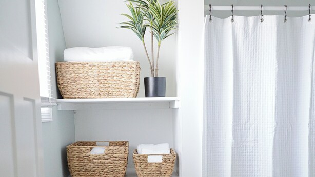 Un baño con estantes blancos que sostienen cestas de mimbre y una planta en maceta.