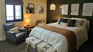 A bedroom with a large bed, a chair, a bedside table with a lamp, a window with blinds, and artwork on the walls. - Ski Paradise (Snowshoe)