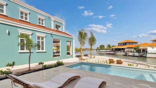 Un espace aménagé au bord de la piscine, avec des chaises longues, une vue dégagée sur l’eau et un bâtiment à la façade bleu clair.