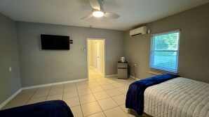 A bedroom with a ceiling fan, a TV, a kitchenette, and a window with blinds. - the nest (Gainesville)