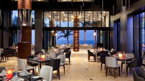 Un restaurant avec des tables dressées pour le service, de grandes fenêtres et une vue sur l’océan.