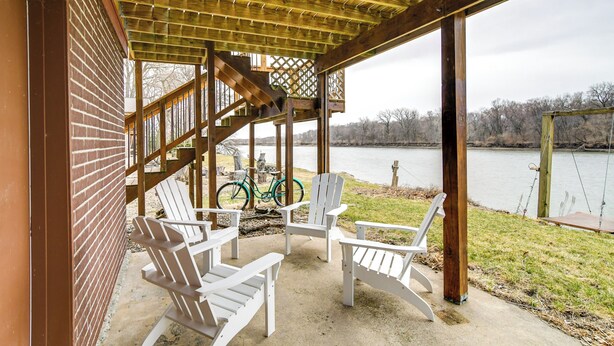 A covered outdoor seating area with Adirondack chairs, a bicycle, and a view of a lake. - Riverfront Cabin Near Dtwn Pella w/ Game Room! (Pella)