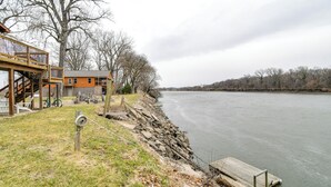 A riverside property with a wooden deck, a stone retaining wall, and a dock extending into the water. - Riverfront Cabin Near Dtwn Pella w/ Game Room! (Pella)
