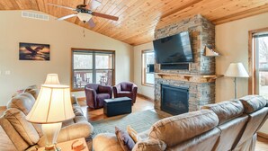 A cozy living room with a stone fireplace, leather sofas, a ceiling fan, and a television mounted above the fireplace. - 6 Mi to Dtwn Pella: Riverfront Cabin w/ Game Room! (Pella)