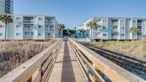 Um calçadão de madeira leva a uma área residencial à beira-mar, com edifícios azul-claros e palmeiras.