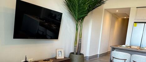 A modern living room with a flat-screen TV mounted on the wall, a wooden console table, a large potted plant, and a dining area with a round table and two chairs.