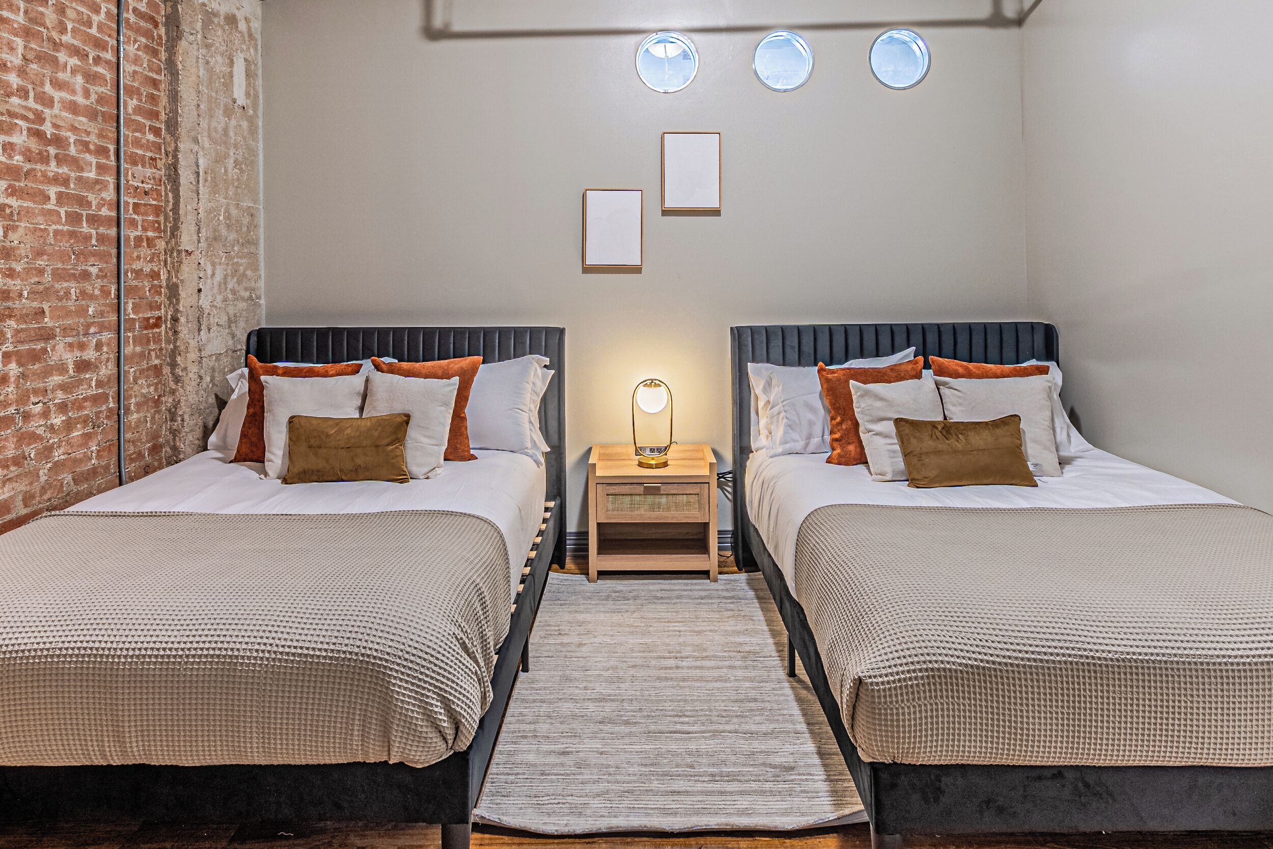 A room with two beds, a brick wall, a small table with a lamp, and three circular ceiling lights.
