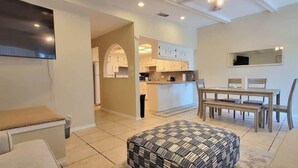 A modern living room with a sofa, ottoman, and a dining area with a table and chairs. There is a kitchen area with cabinets and a microwave.