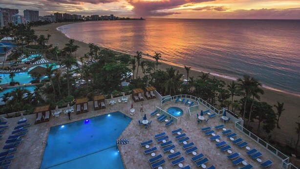A resort with a pool area, lounge chairs, and a beach view during sunset.