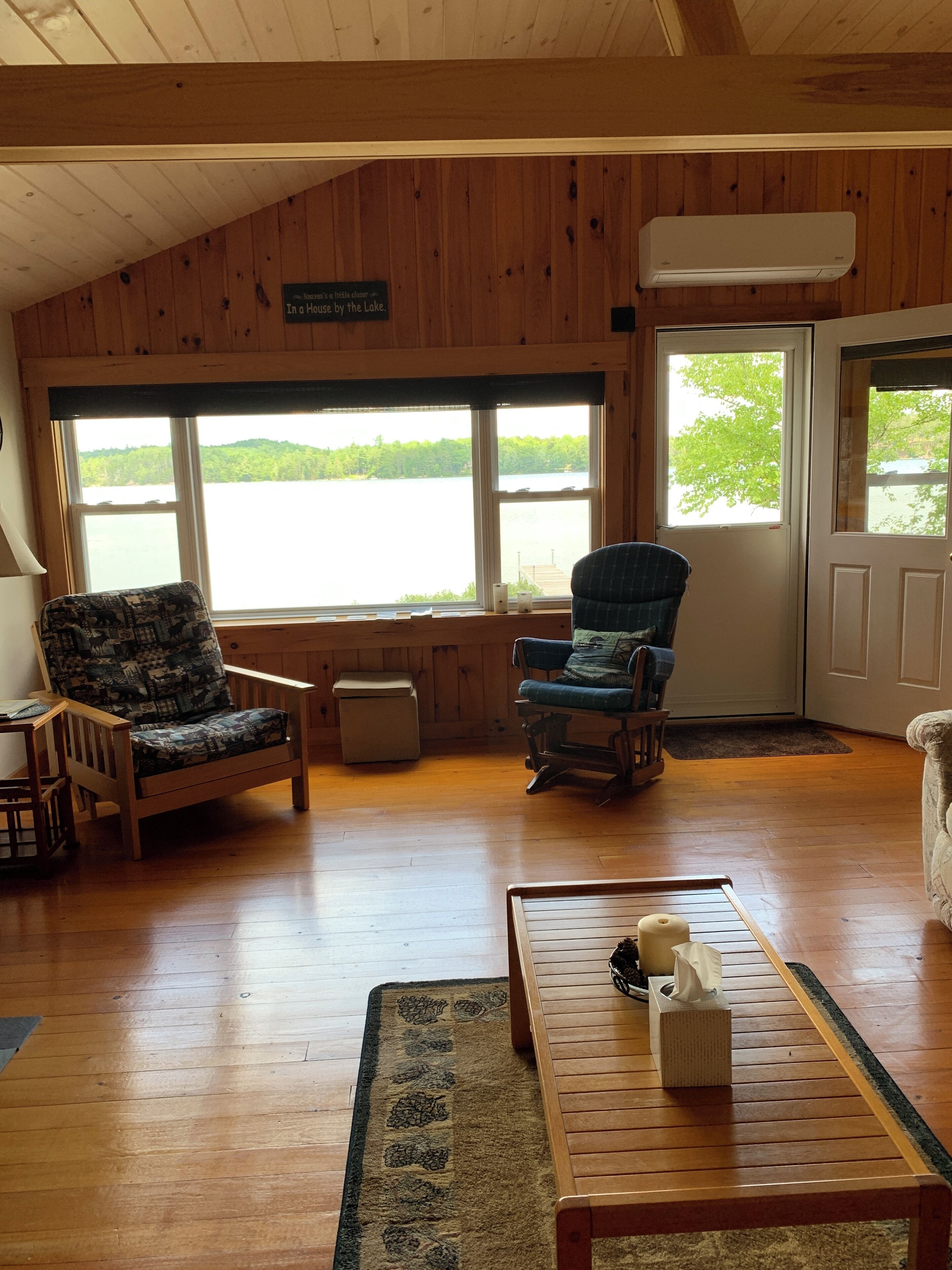 A cozy cabin interior with wooden walls, a large window overlooking a lake, two chairs, a small table with a tissue box, and a rug on the floor.