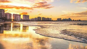 A coastal cityscape at sunset with buildings along the shore and a calm sea reflecting the sky.