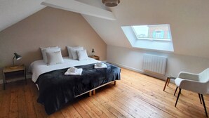 Een slaapkamer met een schuine kap, een dakraam, een bed met witte lakens, een zwarte sprei, een nachtkastje met een lamp en een stoel.