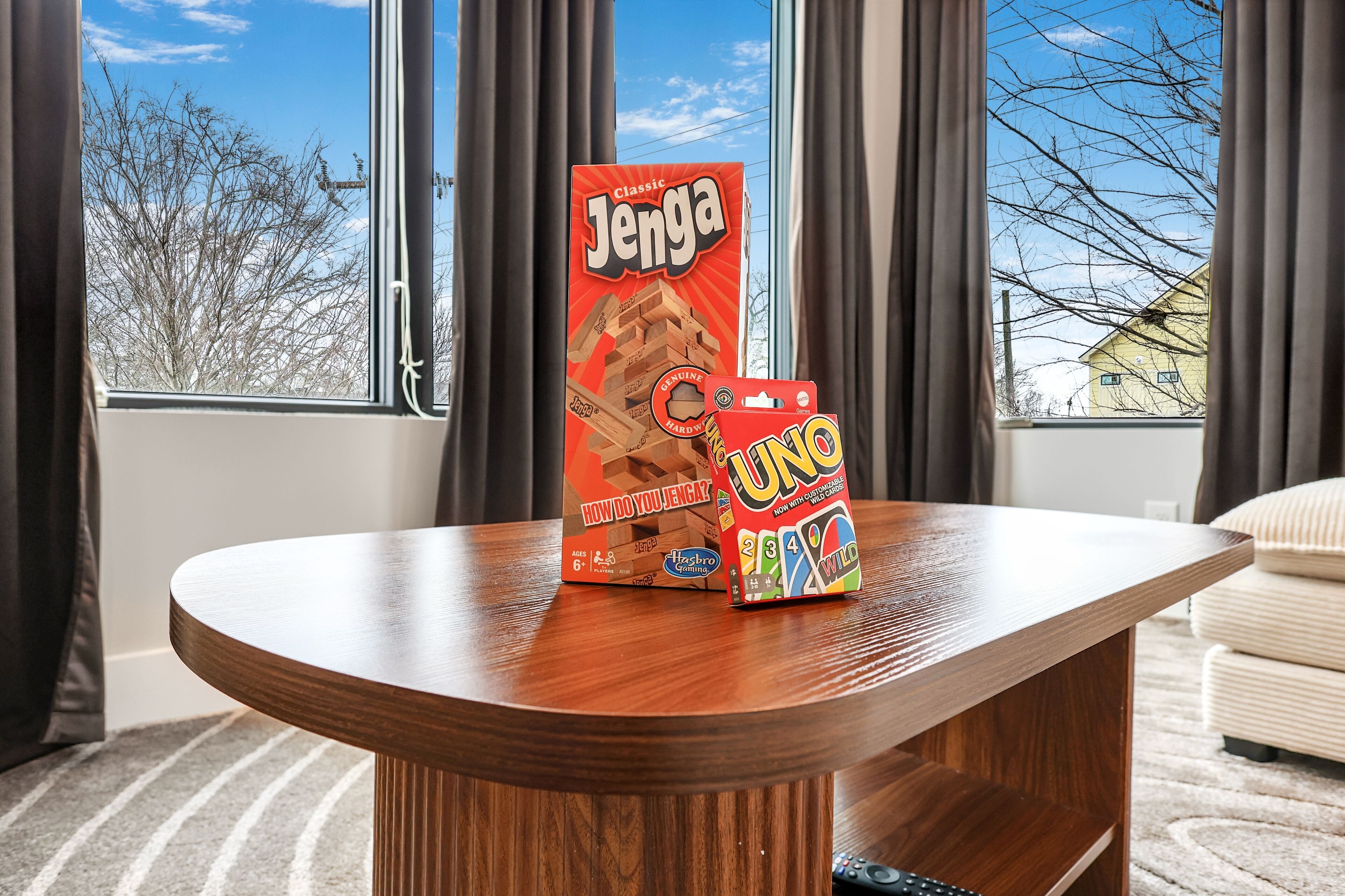 Una mesa de madera con una caja de juego de mesa y otra caja de juego, en una habitación con vistas a árboles y un edificio a través de una ventana.