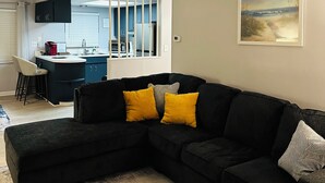 Un salon moderne avec un canapé d’angle, une cuisine en arrière-plan et un tableau accroché au mur.