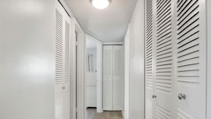 Un couloir étroit aux murs blancs et au plancher en bois, qui mène à une porte dotée d’une poignée blanche.