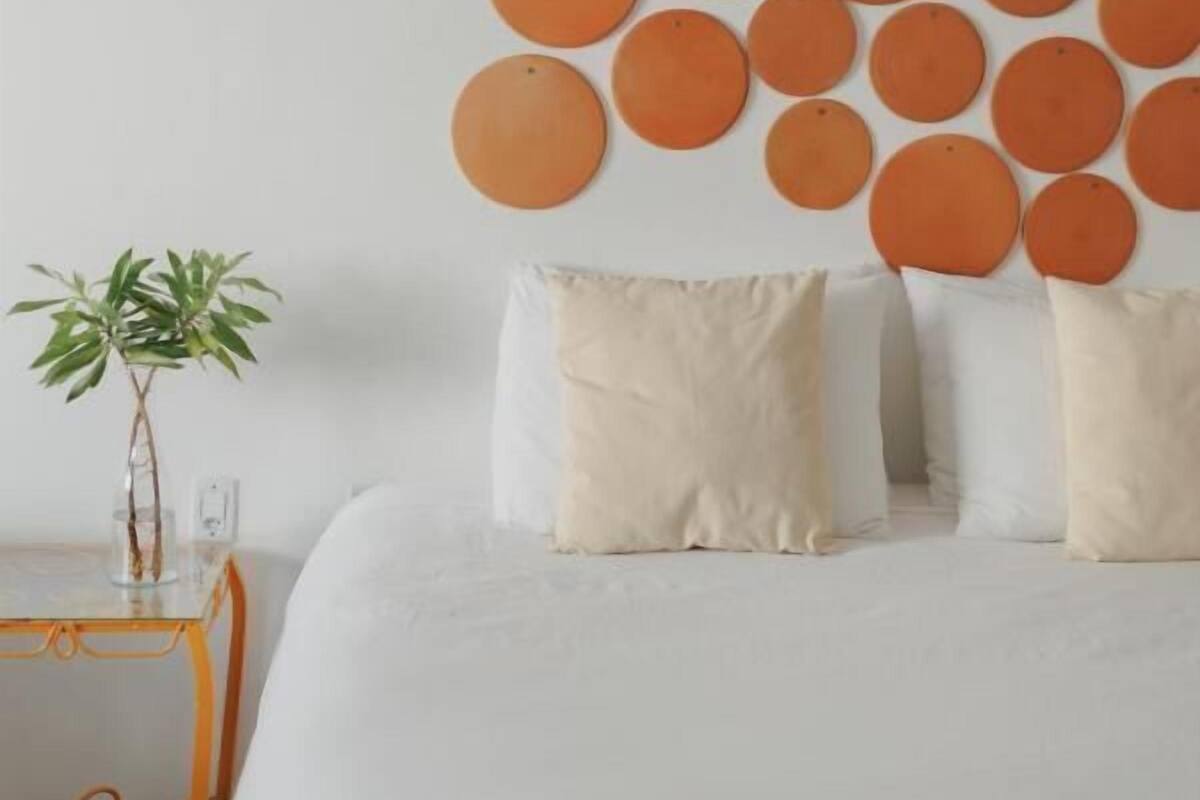 Una cama bien tendida con edredón blanco y dos almohadas, una mesita con una planta, y una pared decorada con adornos naranjas circulares.