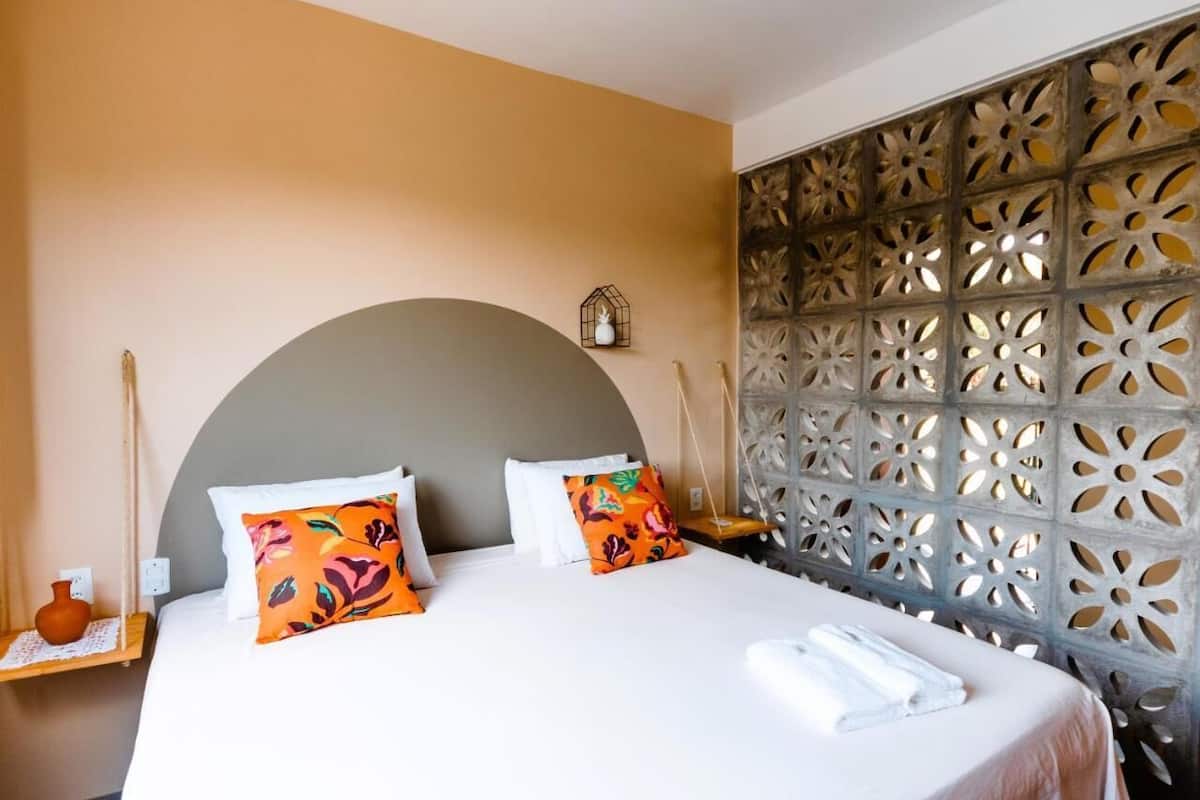 Habitación de hotel con una cama grande, almohadones decorativos, una pared con intrincados diseños tallados y un pequeño estante con un jarrón.