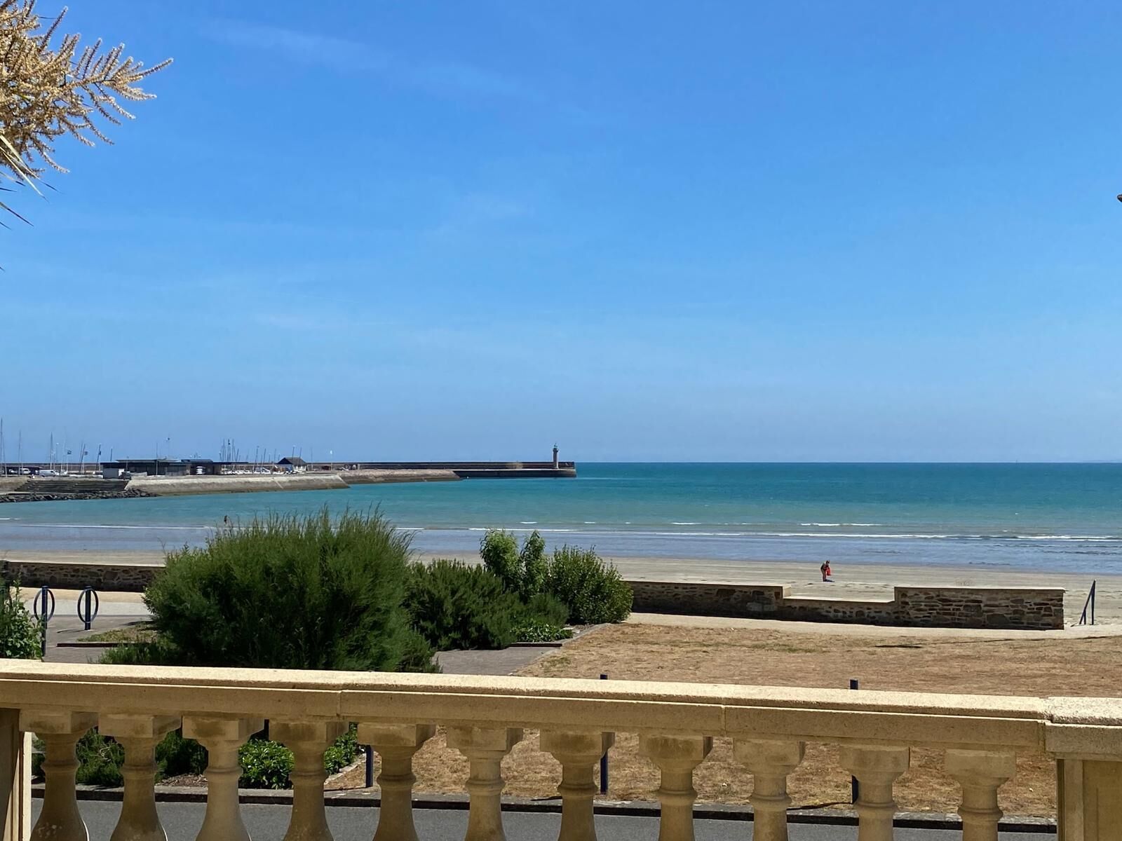 Une vue sur la côte, avec un ciel dégagé, une plage de sable et une jetée en pierre qui s’avance dans l’eau.