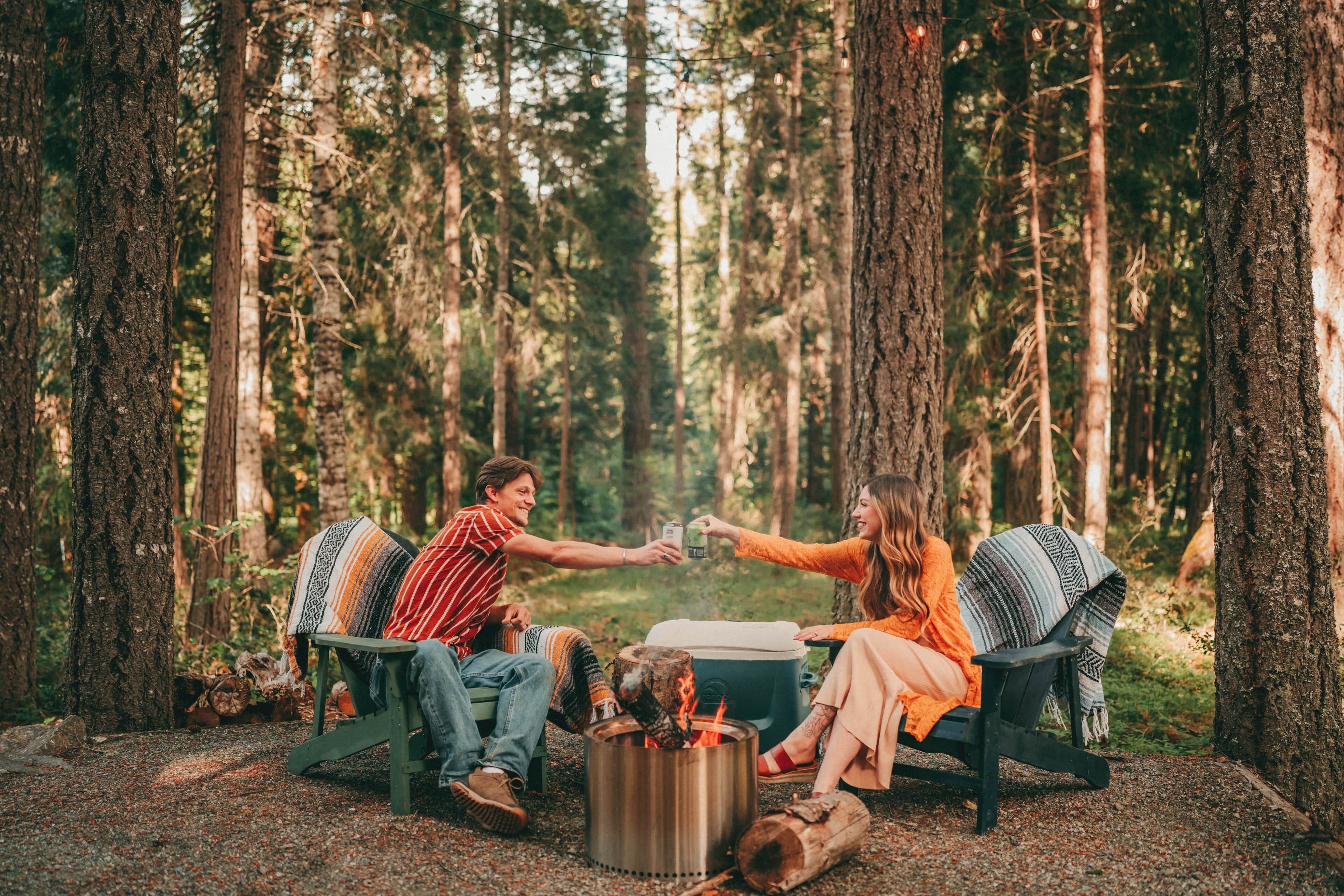 A forest setting with two people sitting on chairs, a fire pit, and a cooler.