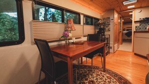 A dining area with a wooden table, chairs, and a rug. There are windows with blinds, a lamp, and a view of trees outside. - Madrone Hideout-Artist Bus Retreat at Azalea Grove Getaway (Douglas County)