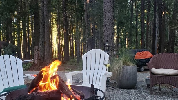 Nuotiopaikka, jonka ympärillä on valkoisia tuoleja ja yksi rottinkituoli. Paikka sijaitsee metsäisellä alueella.