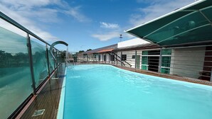 A swimming pool with a wooden deck, glass railings, and a building with a red roof in the background. - Rede Andrade Rio Branco (Rio Branco)