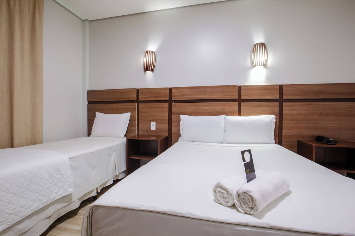 Ett hotellrum med två separata sängar, en stor säng med en handduk och en nyckelring, en sänggavel i trä, sängbord och väggmonterade lampor.