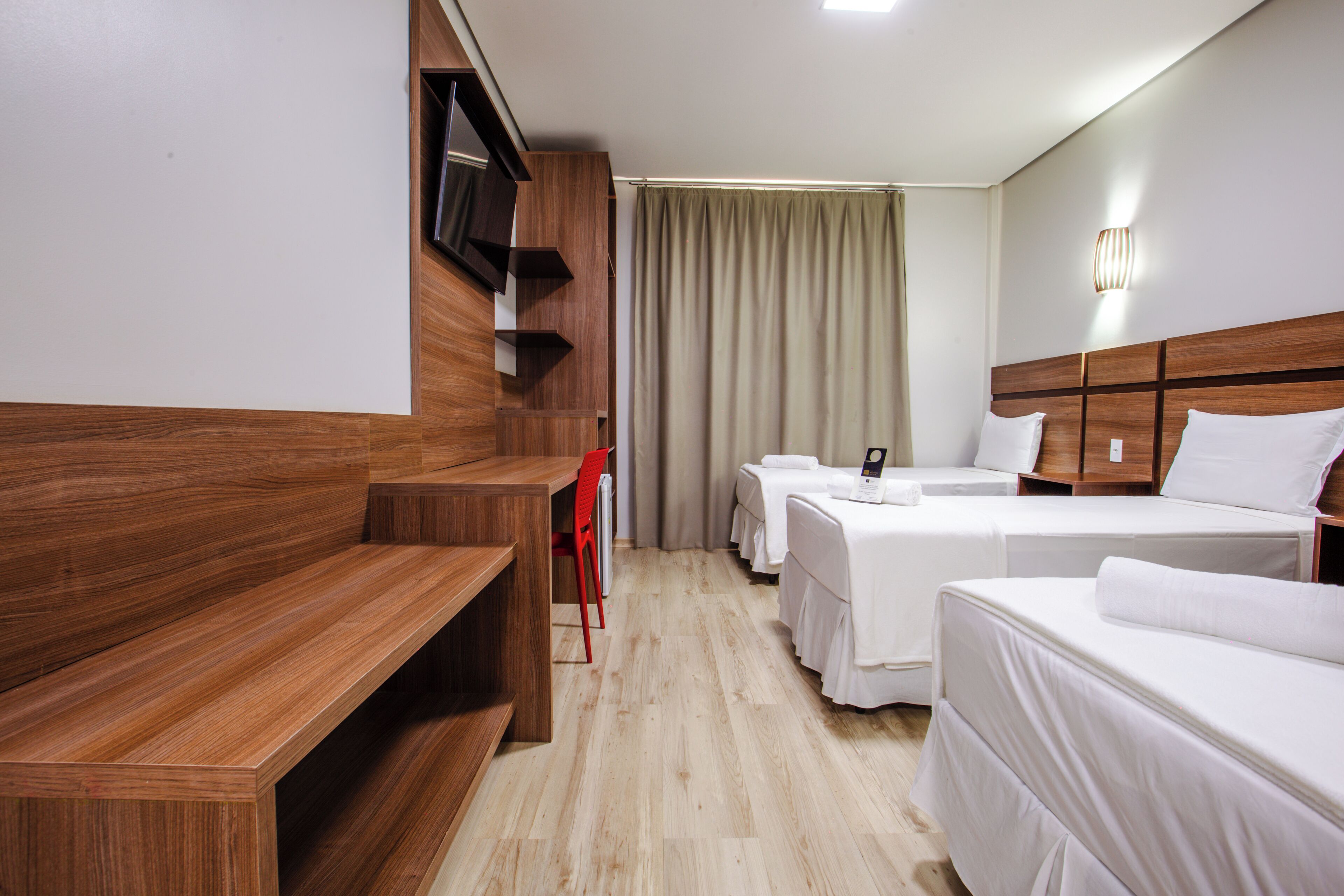 A hotel room with a wooden bench, two single beds, a TV mounted on the wall, and a curtain.