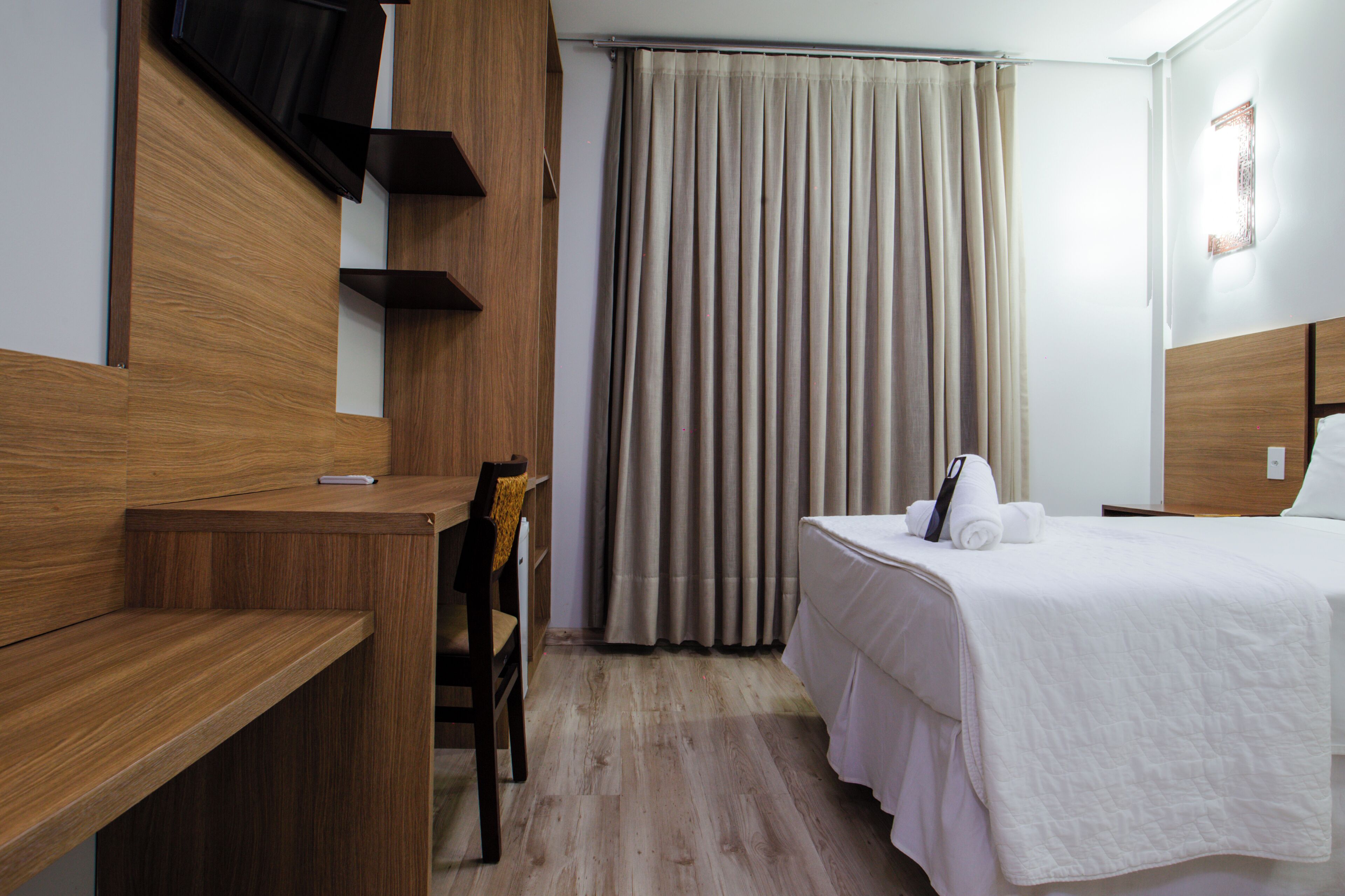 Una habitación de hotel con un escritorio de madera, una cama con cubrecama blanco y un ventanal con cortinas.
