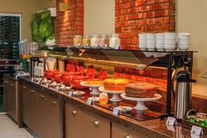 A buffet counter with various food items, a coffee machine, and a trash can. - Rede Andrade Rio Branco (Rio Branco)