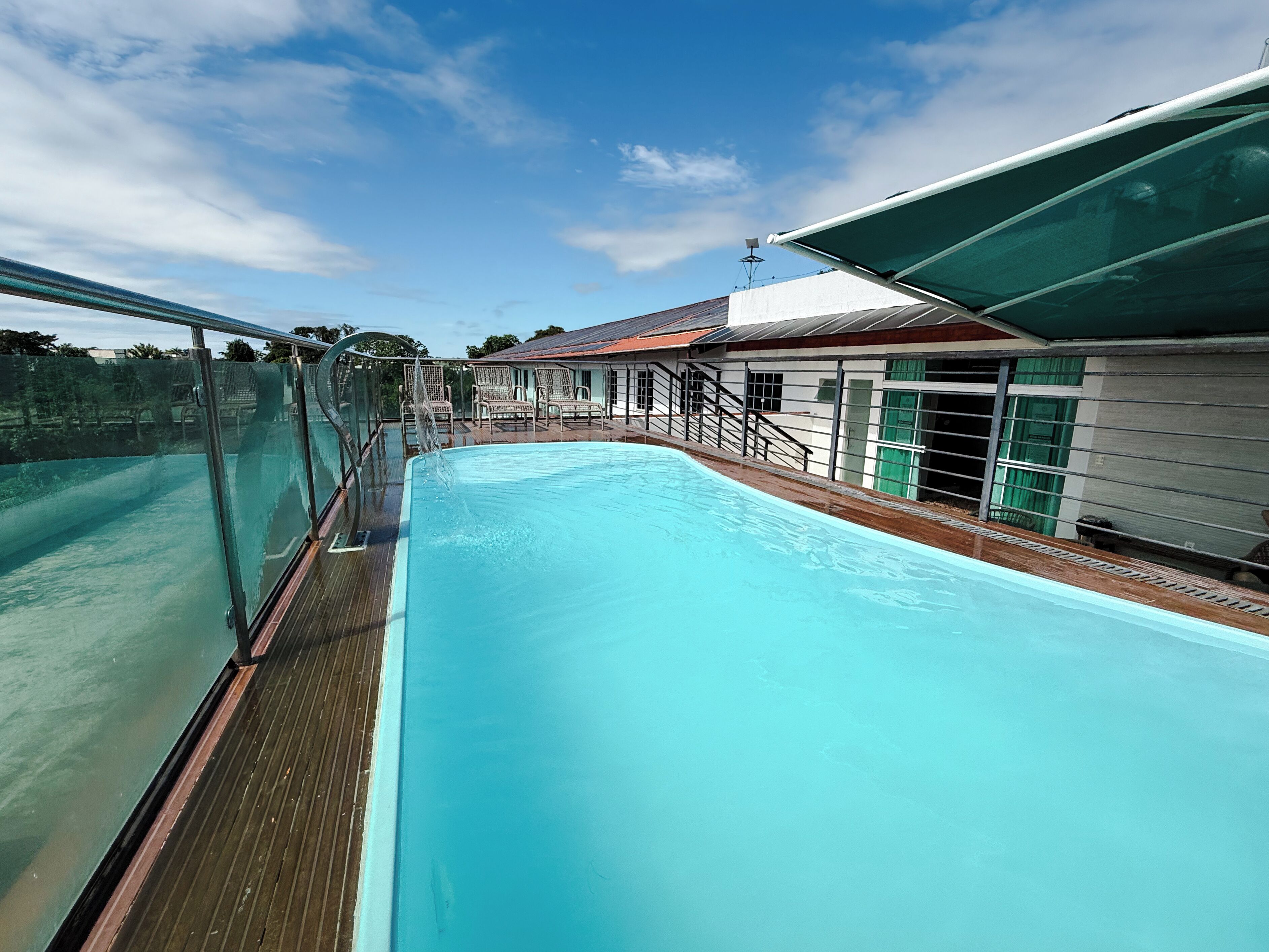 Piscina com deck de madeira, guarda-corpo de vidro e um edifício com telhado vermelho ao fundo.