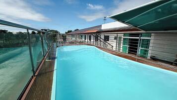 Piscina com deck de madeira, guarda-corpo de vidro e um edifício com telhado vermelho ao fundo.