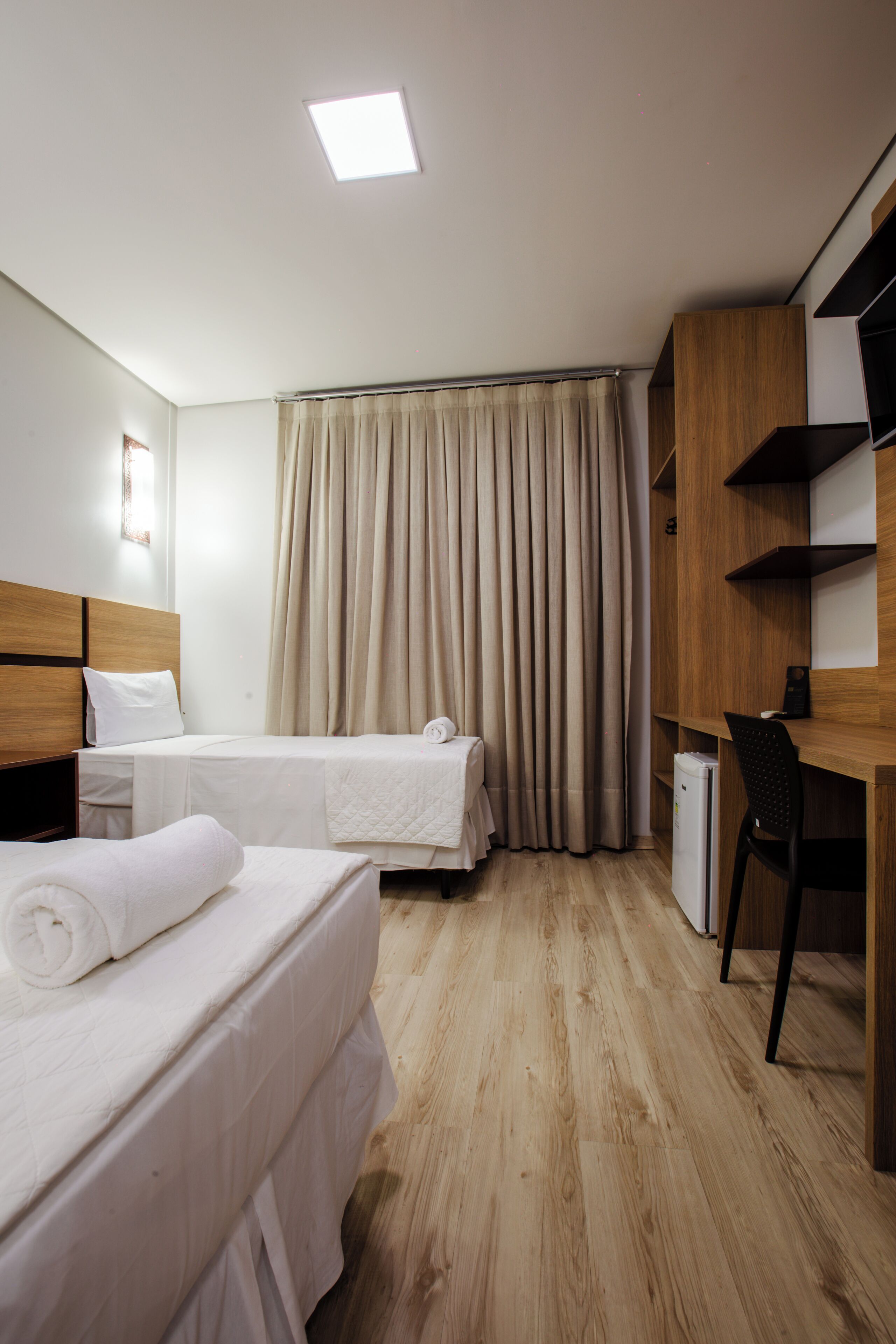 Ein Hotelzimmer mit zwei Betten, einem Schreibtisch, einem Stuhl, einem Kleiderschrank und einem Fenster mit Vorhängen.