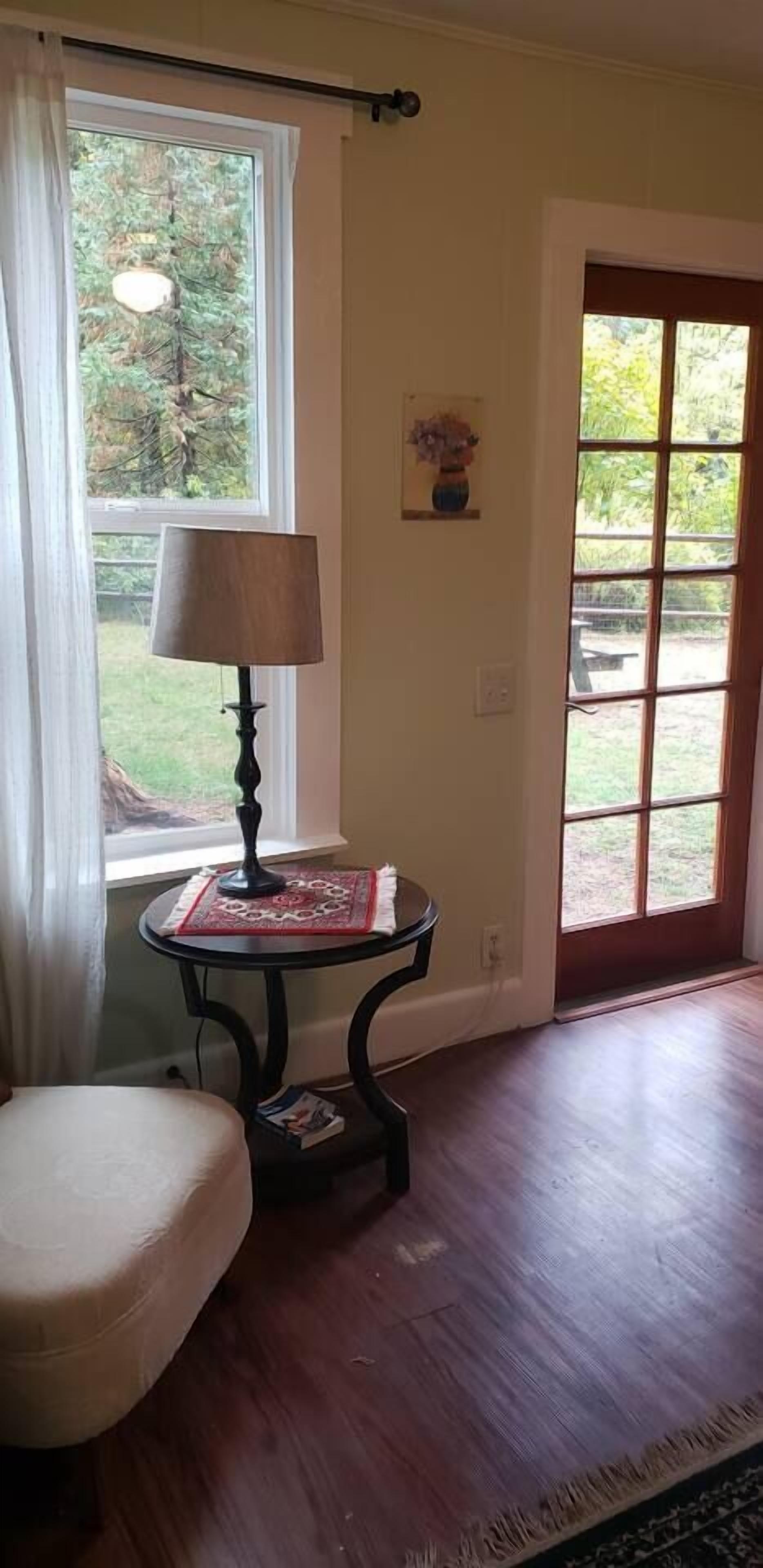A room with a wooden floor, a round table with a lamp, a chair, and a window with a view of greenery.
