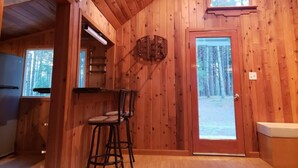 A wooden cabin interior with a kitchen area, a bar stool, a door, and a window overlooking a forest. - Blissful Solitude Cottage (Douglas County)