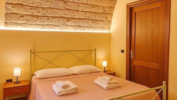 Una cama bien hecha con estructura metálica, dos mesitas de noche con lámparas, una puerta de madera y un techo con textura.