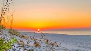 A beach with sand and sparse vegetation, a sunset in the background, and a calm sea. - Sonny Daze~Private Heated Pool~Sleeps 12 (Dauphin Island)