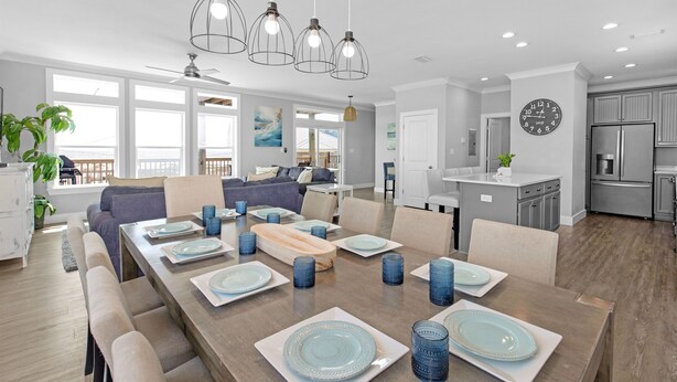 A spacious dining area with a long table set for six, surrounded by chairs. There are pendant lights above the table, and a kitchen area is visible in the background. - Sonny Daze~Private Heated Pool~Sleeps 12 (Dauphin Island)