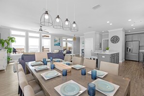 A spacious dining area with a long table set for six, surrounded by chairs. There are pendant lights above the table, and a kitchen area is visible in the background. - Sonny Daze~Private Heated Pool~Sleeps 12 (Dauphin Island)
