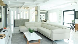 Una sala de estar moderna con un amplio sofá seccional en forma de L, una mesa de centro blanca y una cocina al fondo.