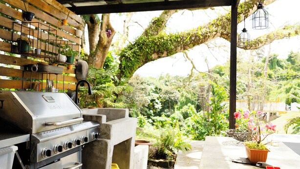 Una parrilla de barbacoa bajo una pérgola de madera con plantas en macetas e iluminación colgante.