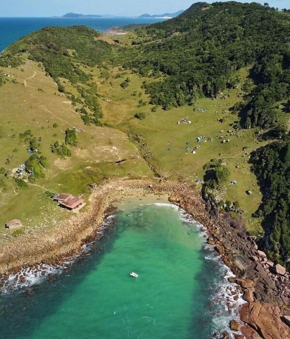 An aerial view of a coastal area with a clear beach, rocky shoreline, and dense greenery. - Casa de Frente pro Mar com Piscina (Palhoca)