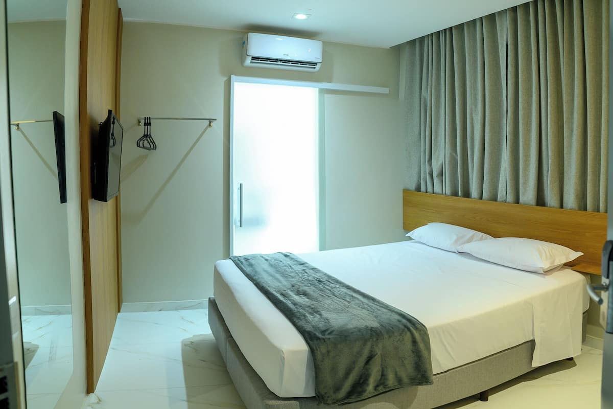 A modern hotel room with a large bed, a flat-screen TV mounted on the wall, a ceiling-mounted air conditioner, and a window with curtains.