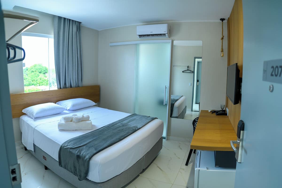 A hotel room with a bed, a desk, a TV, and an air conditioning unit.