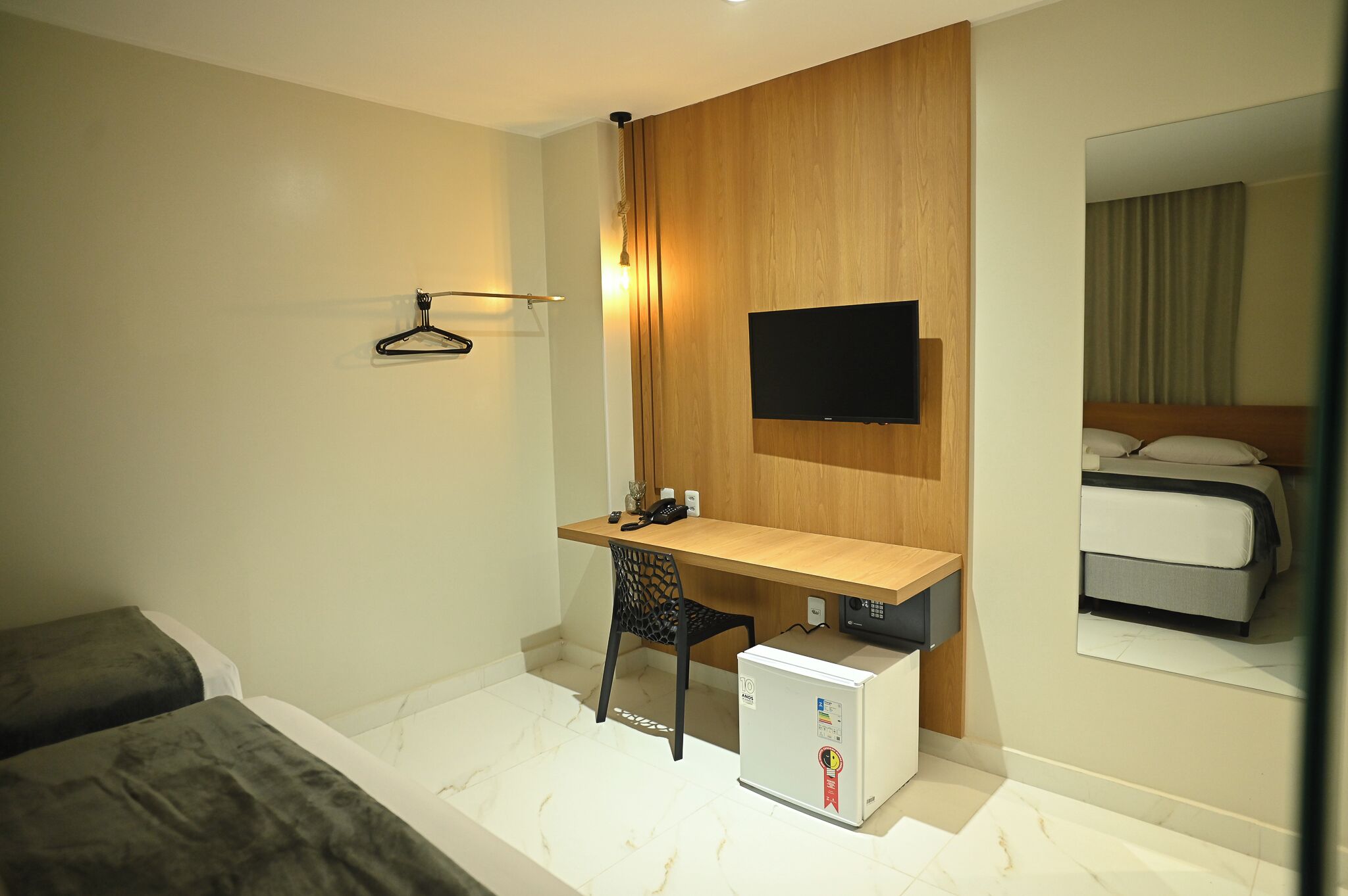 Una habitación de hotel con un escritorio de madera, una silla, un televisor montado en la pared, una cama con cabecera y un refrigerador pequeño.
