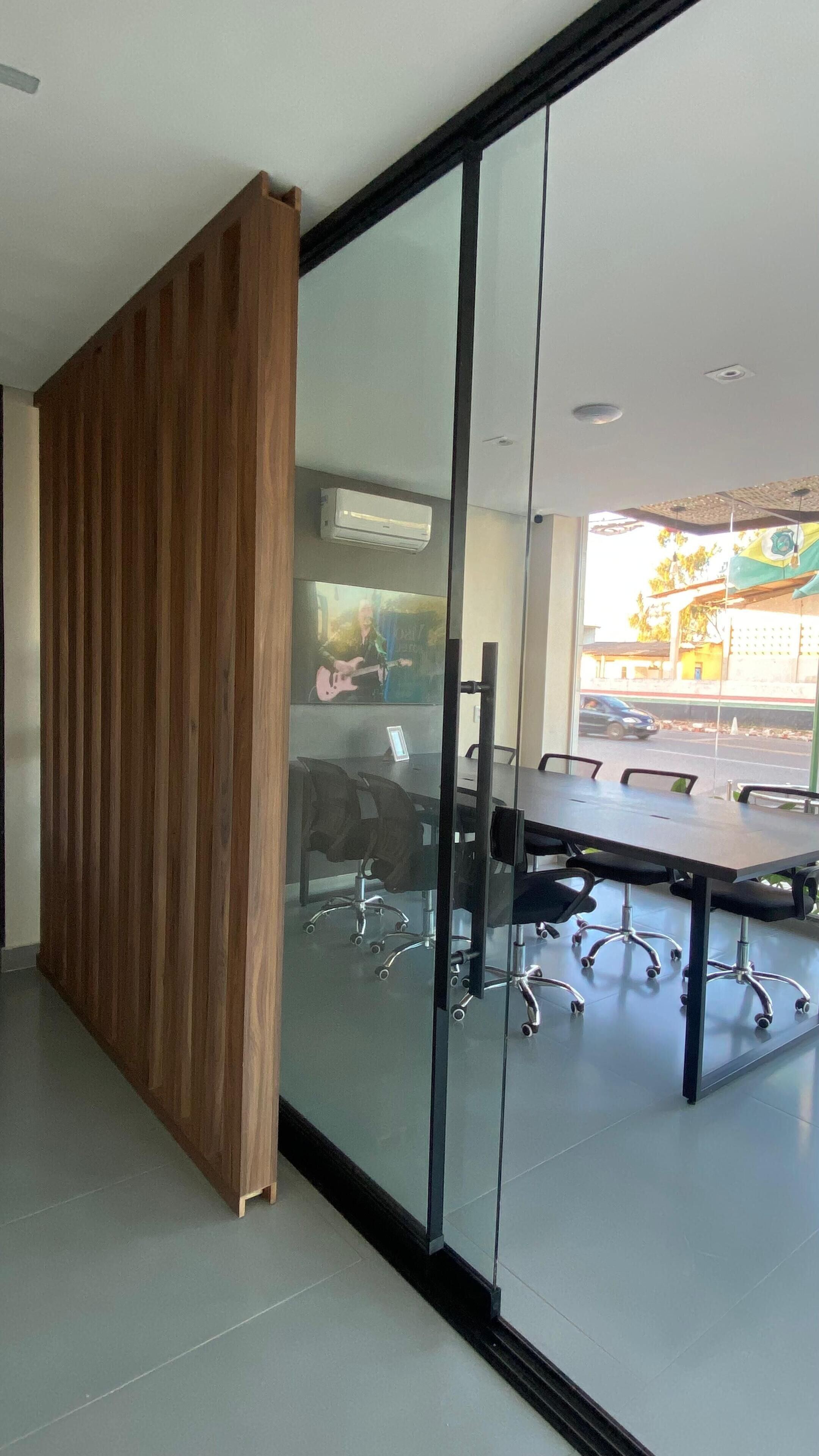 A modern meeting room with a glass partition, a wooden panel, and a large table with office chairs.