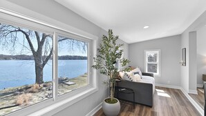 Un dormitorio moderno con una ventana grande que da a un lago, un sofá gris y una planta en maceta.