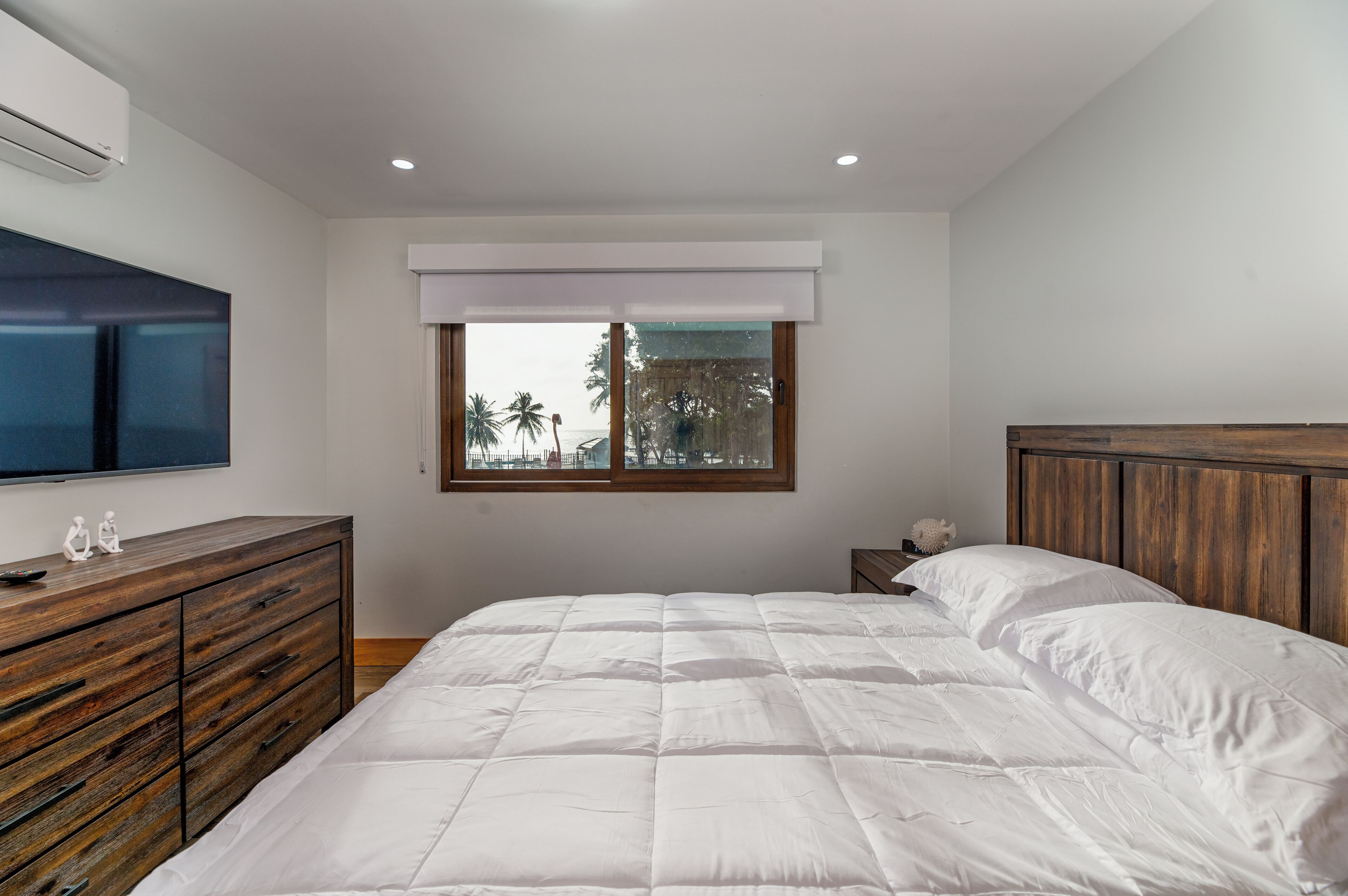 A bedroom with a large bed, wooden dresser, TV, and a window with a view of palm trees.