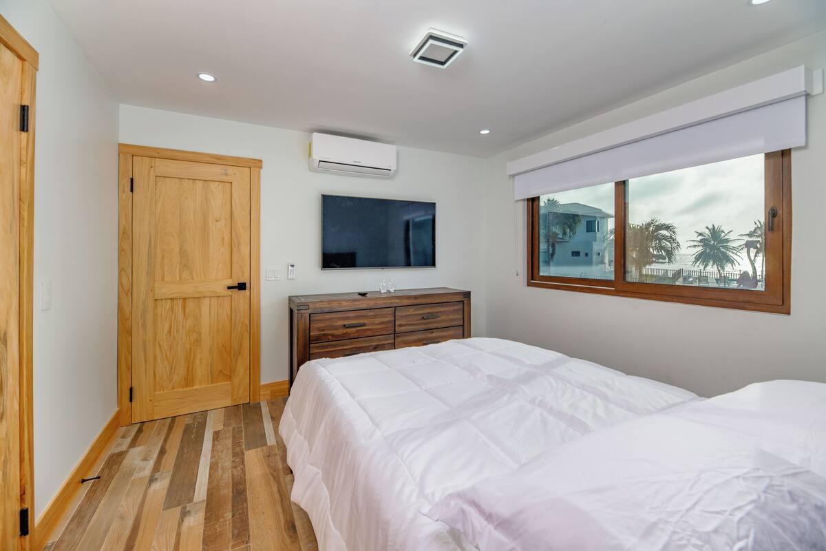 A bedroom with a wooden wardrobe, a TV mounted on the wall, a bed with white bedding, and a window with a view of palm trees.