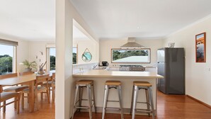 A modern kitchen with a central island, bar stools, a refrigerator, and a window overlooking greenery. - Bodhi's Reef | Coastal Retreat (Lake Tyers Beach)